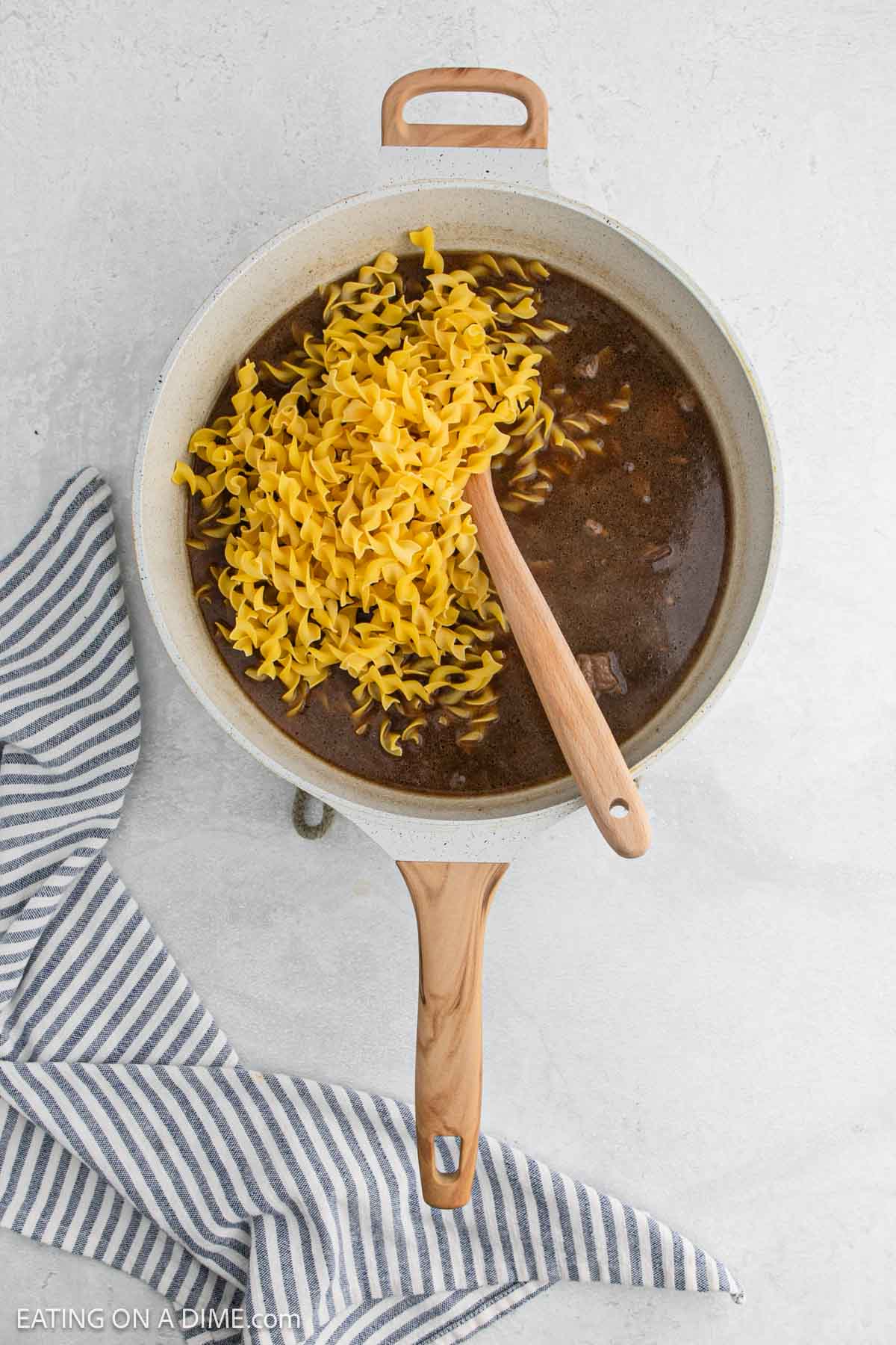 A pot of French Onion Beef stew with uncooked egg noodles on top and a wooden spoon resting inside, placed on a light surface next to a striped cloth.