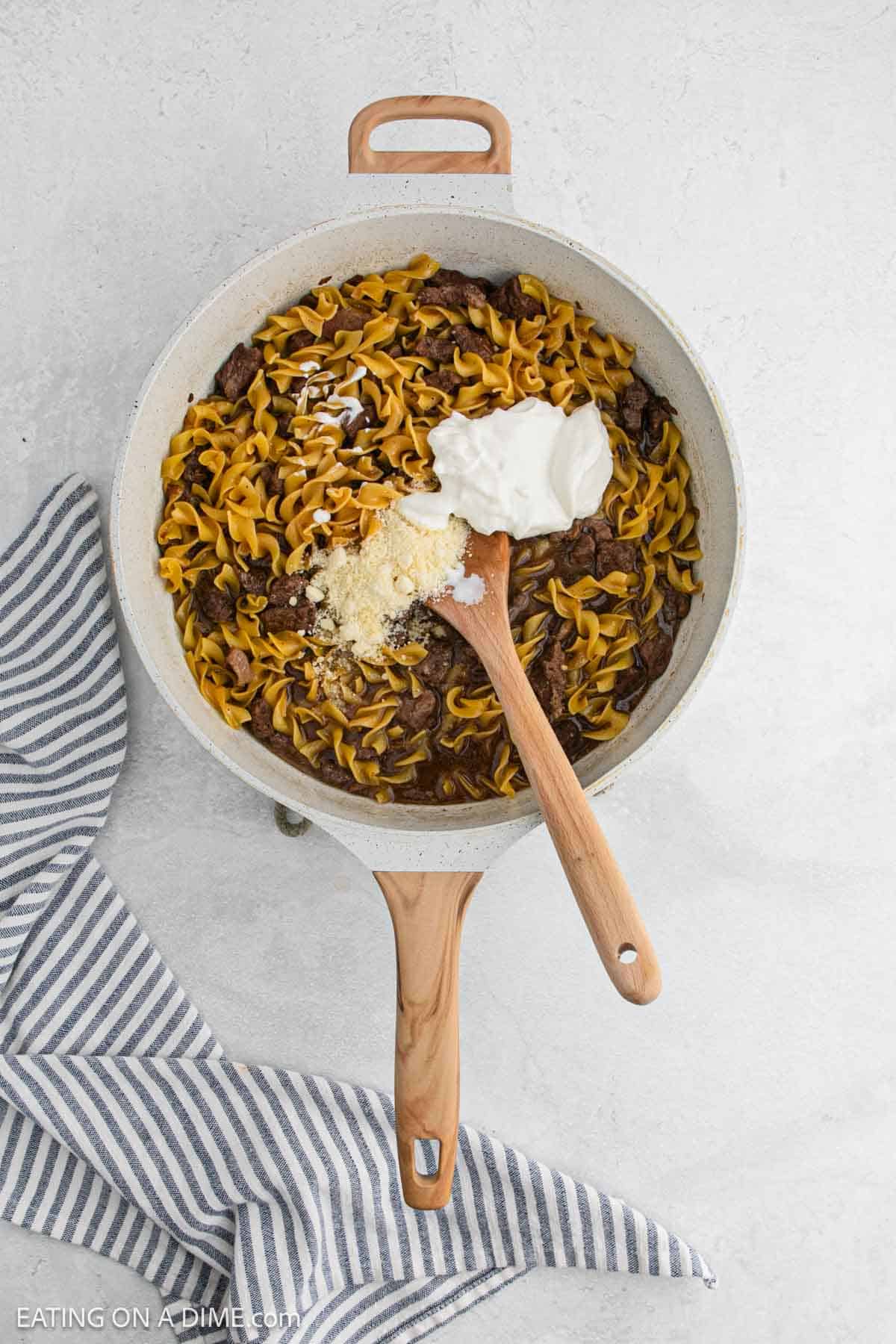Overhead view of a skillet with French Onion Beef and noodles, topped with sour cream and grated cheese. A wooden spoon rests inside the pan, which sits on a light surface beside a striped towel.