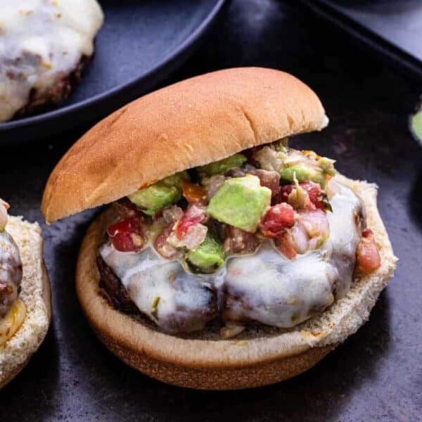 A hamburger with melted cheese, topped with diced avocado, tomato, and onion mixture, served in a bun on a dark surface—perfect for adding variety to your Meal Plan 61.