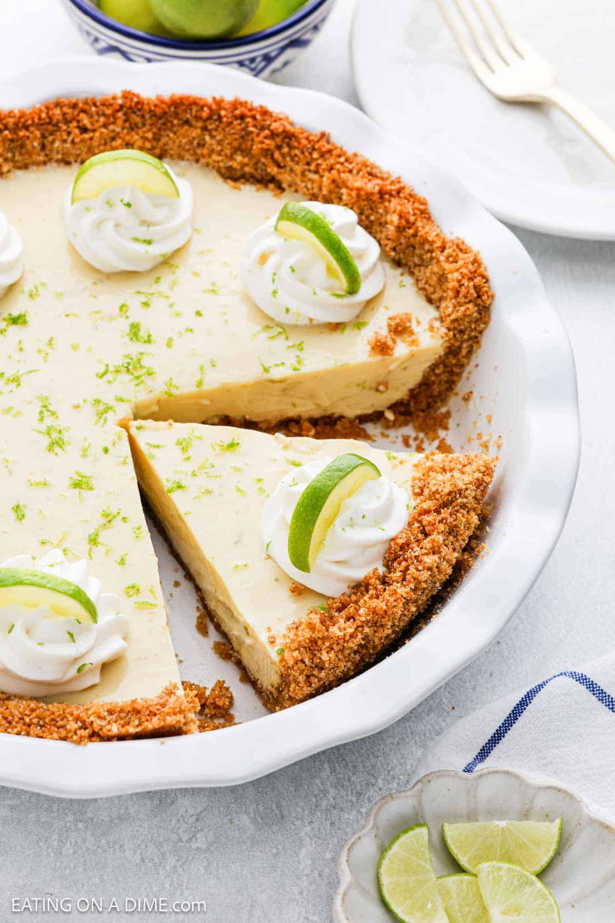 A Key Lime Pie with a graham cracker crust sits in a white pie dish, topped with whipped cream and lime wedges. One slice is missing, and nearby are plates, a fork, and fresh lime slices.