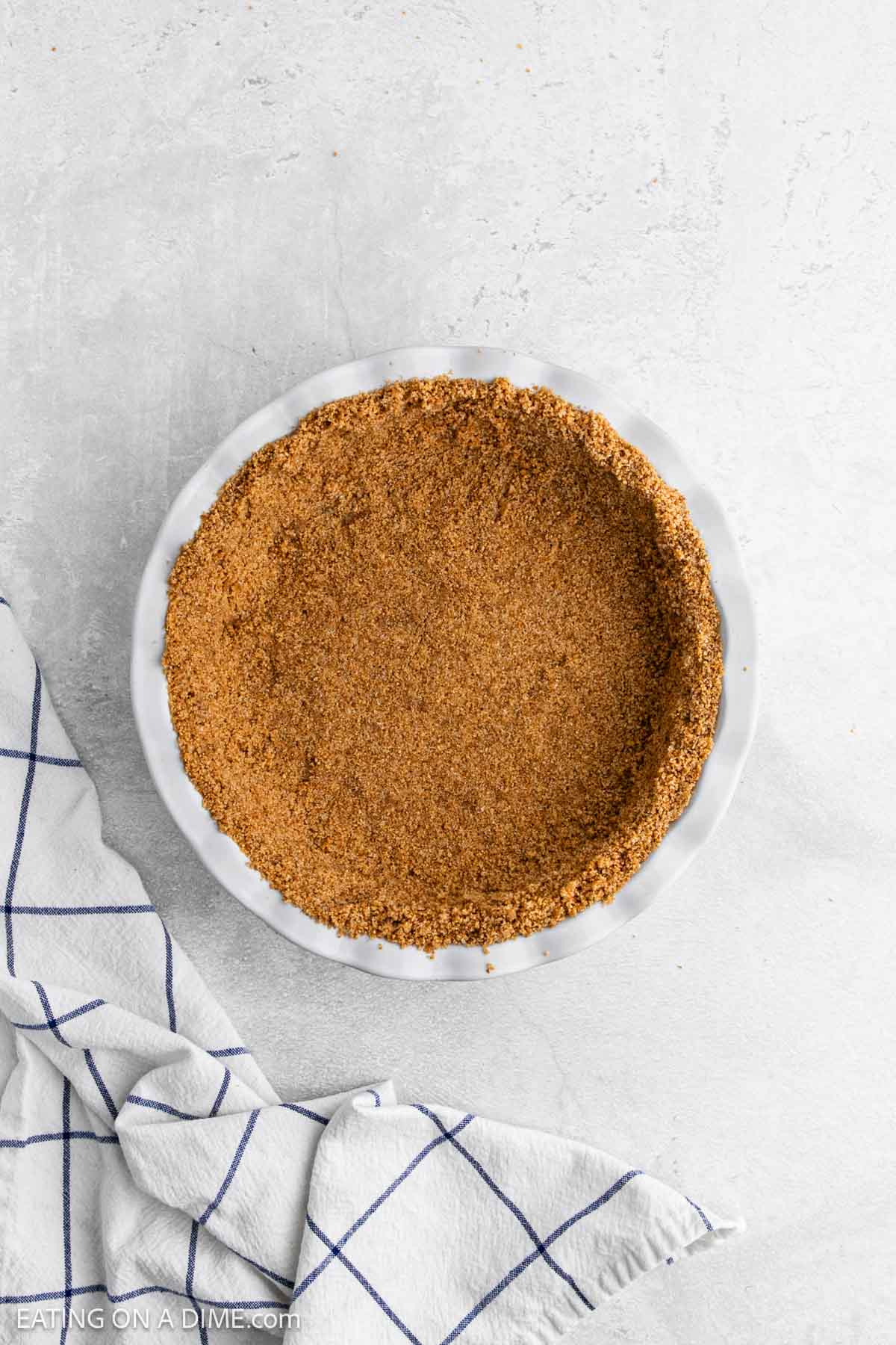 A graham cracker pie crust, perfect for Key Lime Pie, sits in a white pie dish on a light gray surface beside a white kitchen towel with blue stripes.