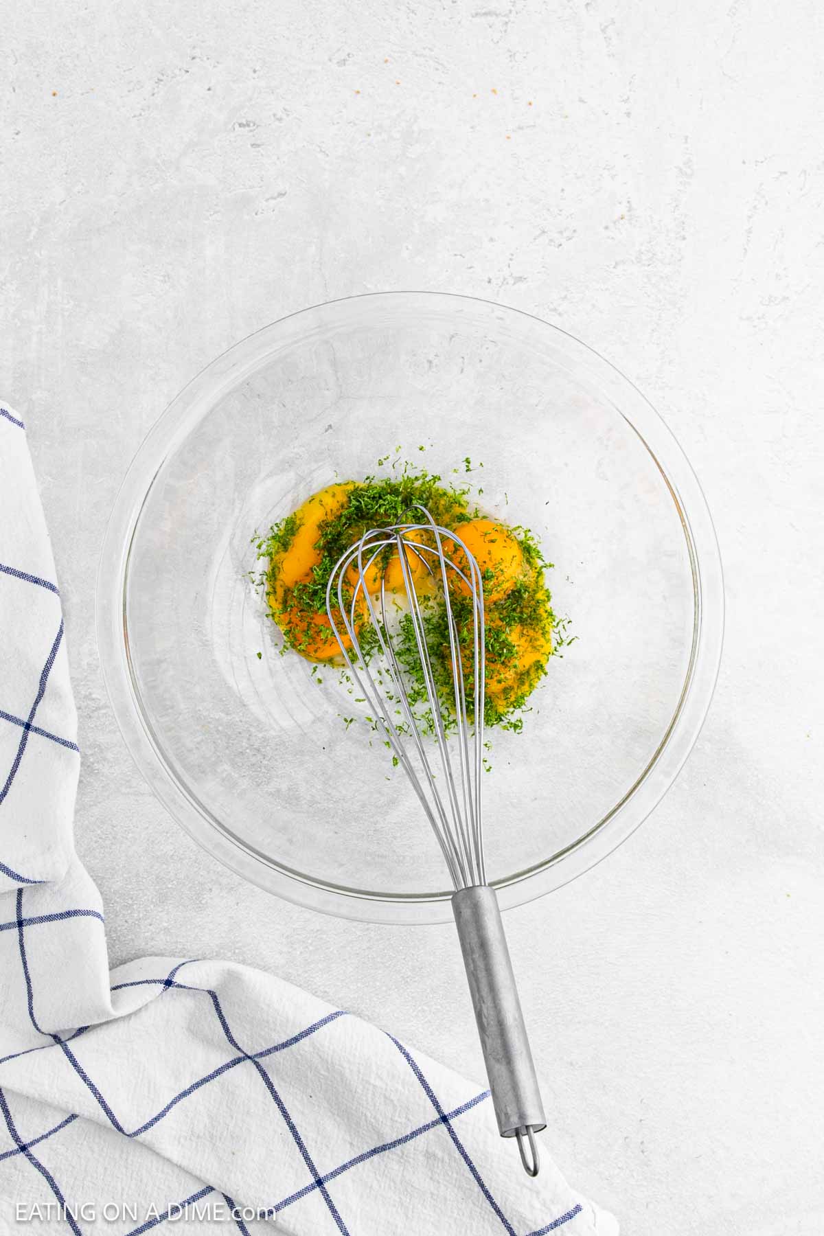 A glass bowl with cracked eggs and chopped herbs, with a metal whisk resting inside. The bowl sits on a light surface next to a white towel with blue grid lines, ready for the next step in making Key Lime Pie.