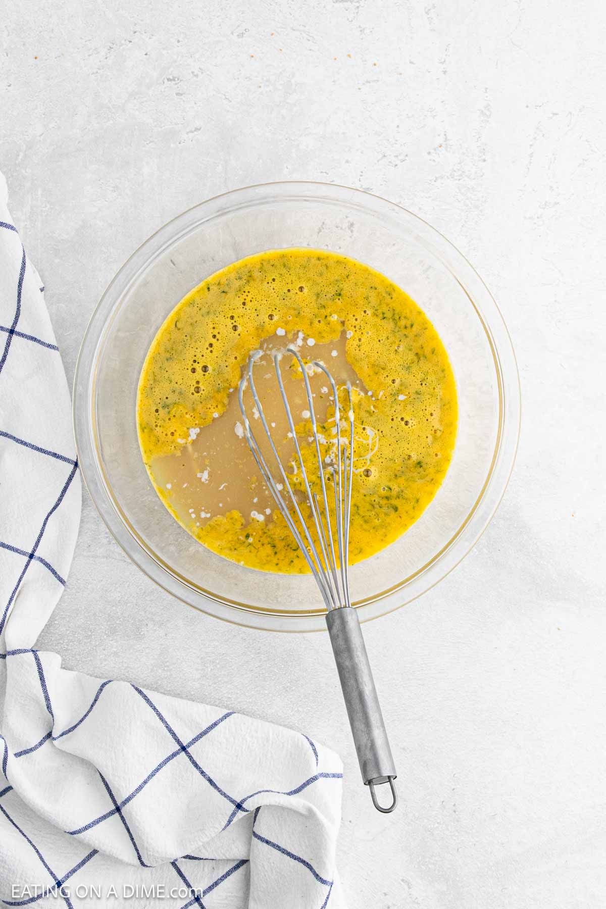 A glass bowl filled with beaten eggs and seasoning, perfect for making Key Lime Pie, sits on a light surface. A metal whisk rests inside the bowl, and a white kitchen towel with blue stripes is nearby.
