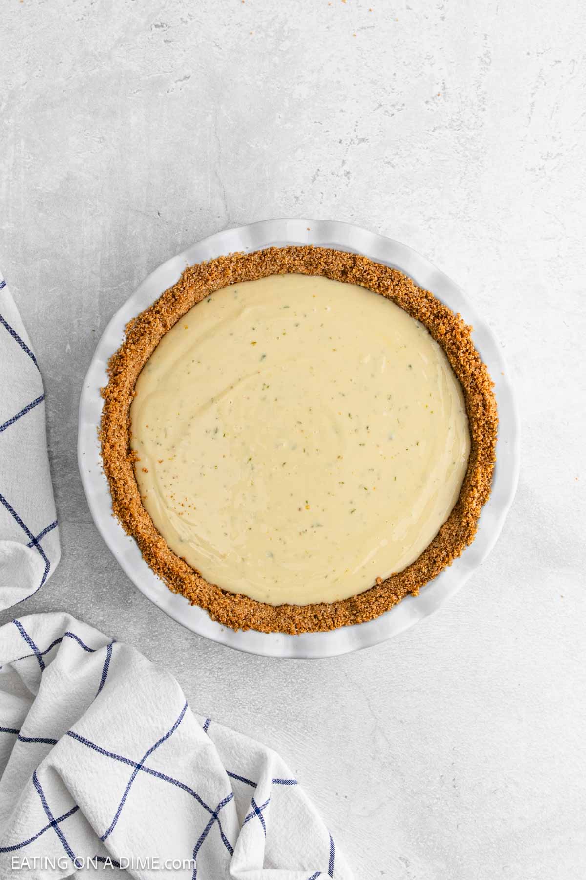 A creamy Key Lime Pie with a graham cracker crust sits in a white pie dish on a light gray surface. A white kitchen towel with blue stripes is partially visible next to the pie.