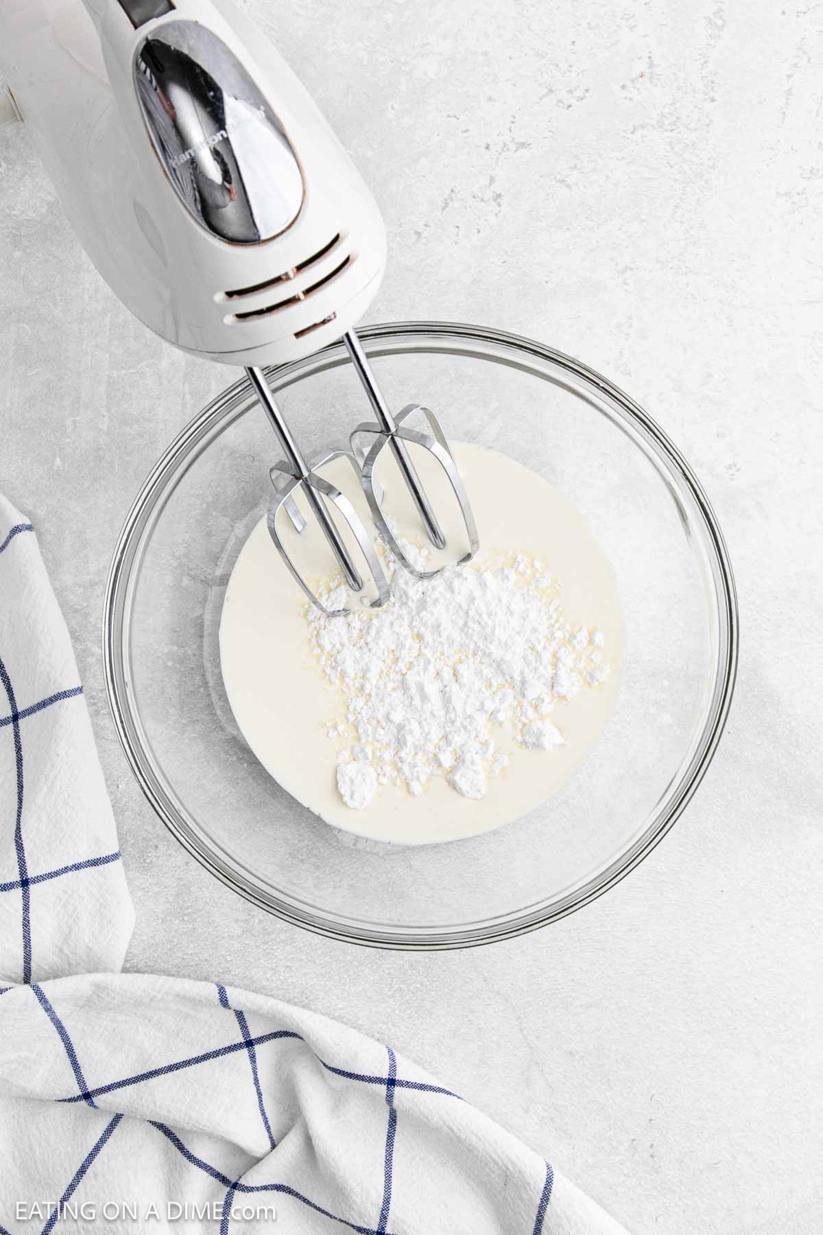 A hand mixer with beaters is positioned over a glass bowl containing cream and powdered sugar, ready to be mixed for a delicious Key Lime Pie. A blue and white checkered towel is partially visible on the side.