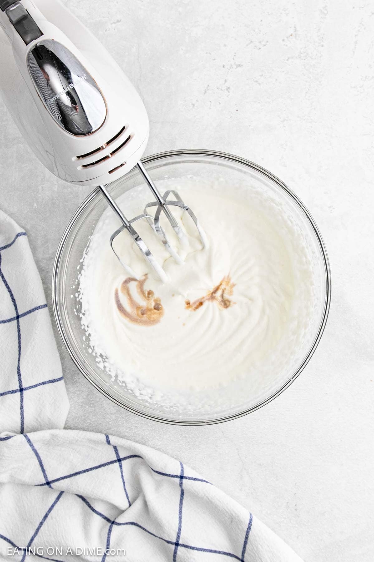 A hand mixer whipping cream and vanilla for a Key Lime Pie in a glass bowl on a light countertop, with a white and blue checkered towel nearby.