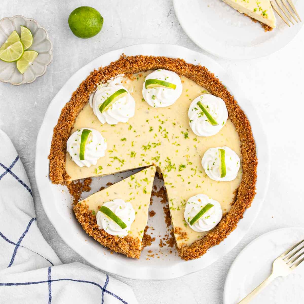 A classic Key Lime Pie with a graham cracker crust, topped with whipped cream and lime slices. One slice is missing, with a lime and wedges nearby. A blue-and-white towel and plates with forks complete the scene.