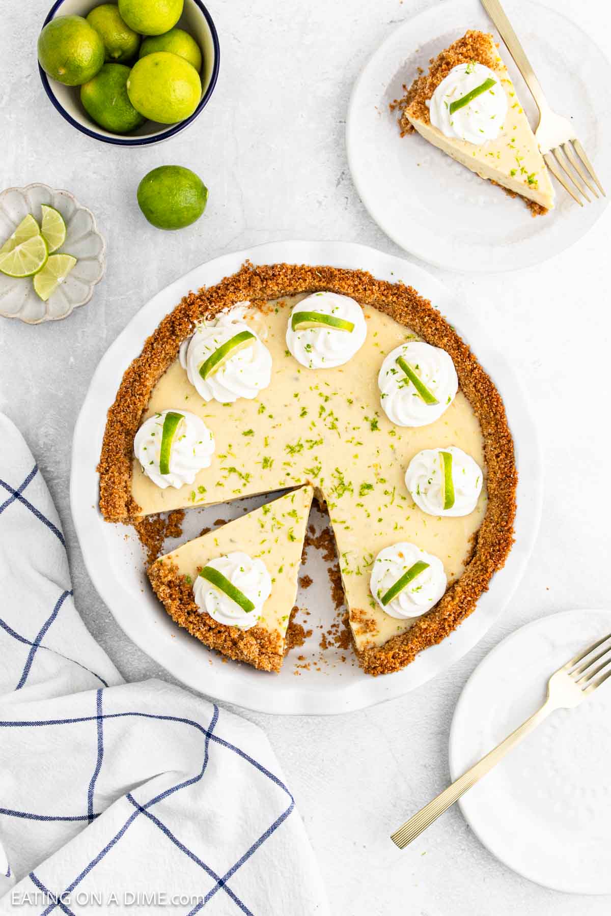 A classic Key Lime Pie with a graham cracker crust, topped with whipped cream and lime slices, sits on a white plate with one slice removed. Slices of this tangy dessert are served nearby, and fresh limes are in a bowl beside the pie.