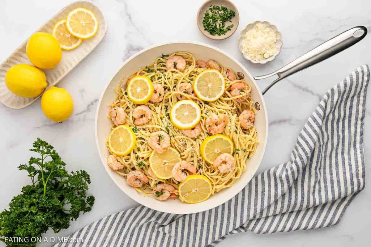 A skillet filled with Lemon Garlic Shrimp Pasta garnished with lemon slices and herbs sits on a marble surface, surrounded by fresh lemons, parsley, cheese, and a striped kitchen towel.