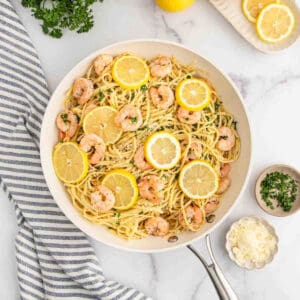 A skillet of Lemon Garlic Shrimp Pasta garnished with lemon slices and parsley, surrounded by a striped towel, grated cheese, chopped herbs, and lemon slices on a marble surface.