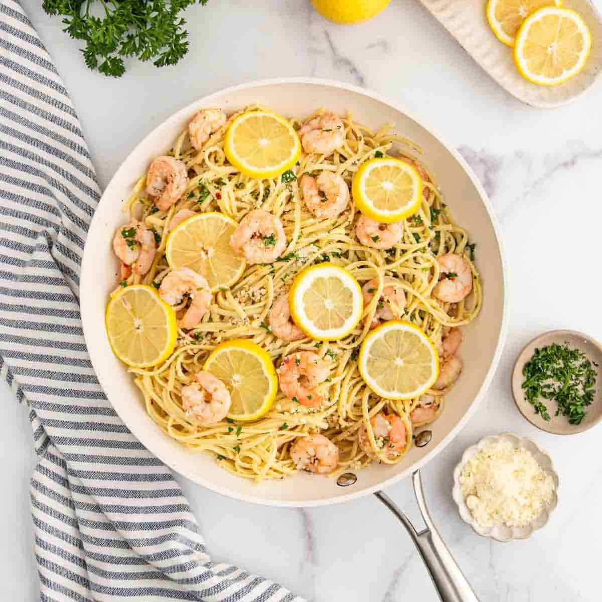 A skillet of Lemon Garlic Shrimp Pasta garnished with lemon slices and parsley, surrounded by a striped towel, grated cheese, chopped herbs, and lemon slices on a marble surface.