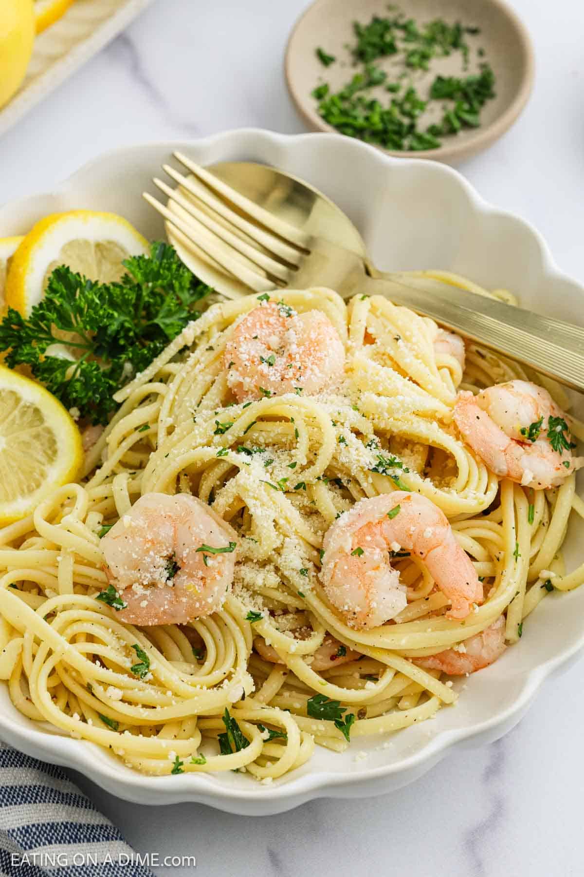 A bowl of Lemon Garlic Shrimp Pasta features linguine topped with shrimp, grated Parmesan cheese, and chopped parsley, garnished with lemon slices and parsley sprigs. A fork and spoon rest on the side of the bowl.