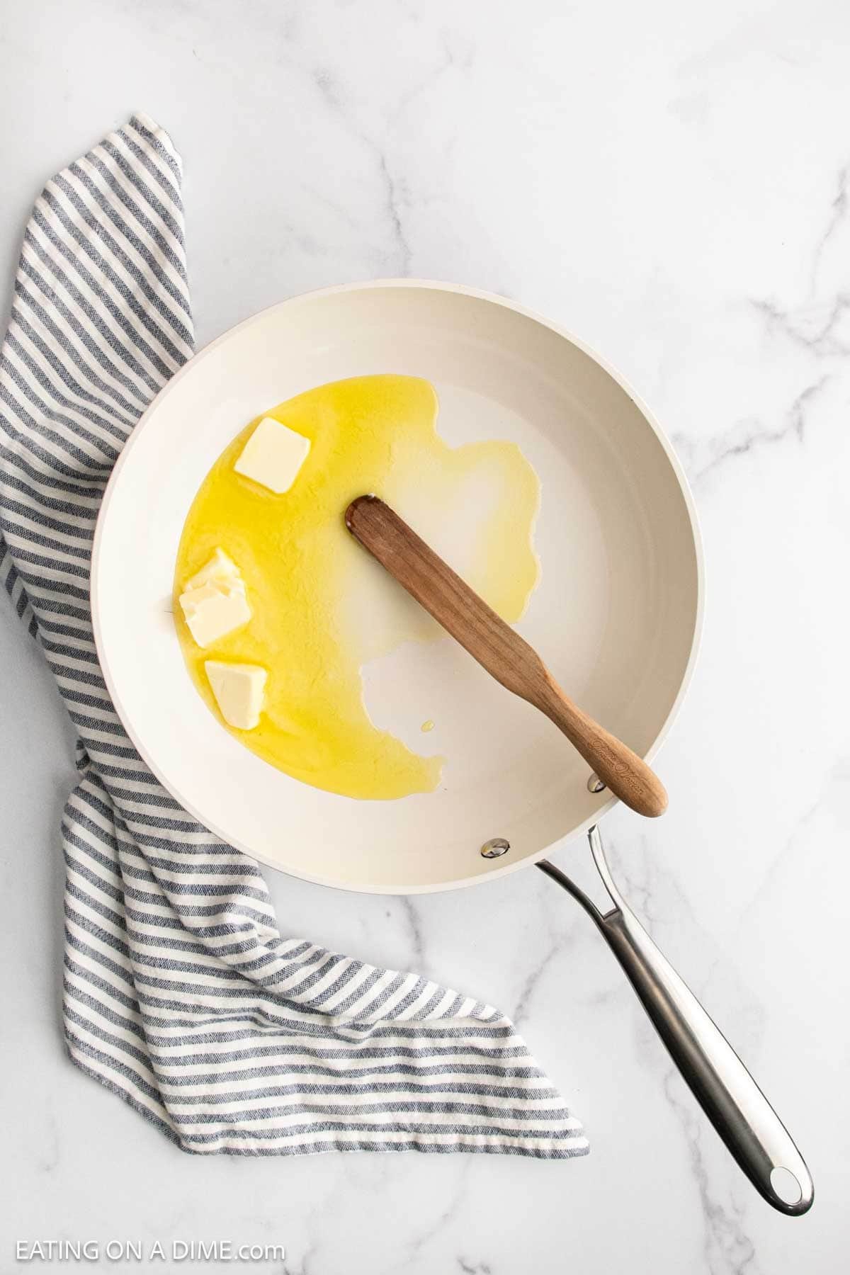 A white skillet with melting butter and a wooden spatula inside, ready to start Lemon Garlic Shrimp Pasta, rests on a striped gray and white kitchen towel on a marble countertop.