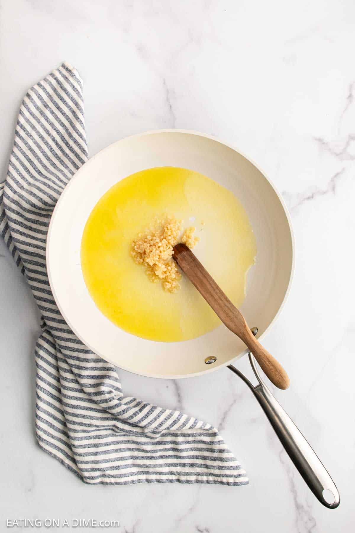 A white skillet with melted butter and minced garlic, perfect for starting Lemon Garlic Shrimp Pasta, sits on a white marble surface. A wooden spatula rests in the pan, and a gray-and-white striped kitchen towel lies underneath.
