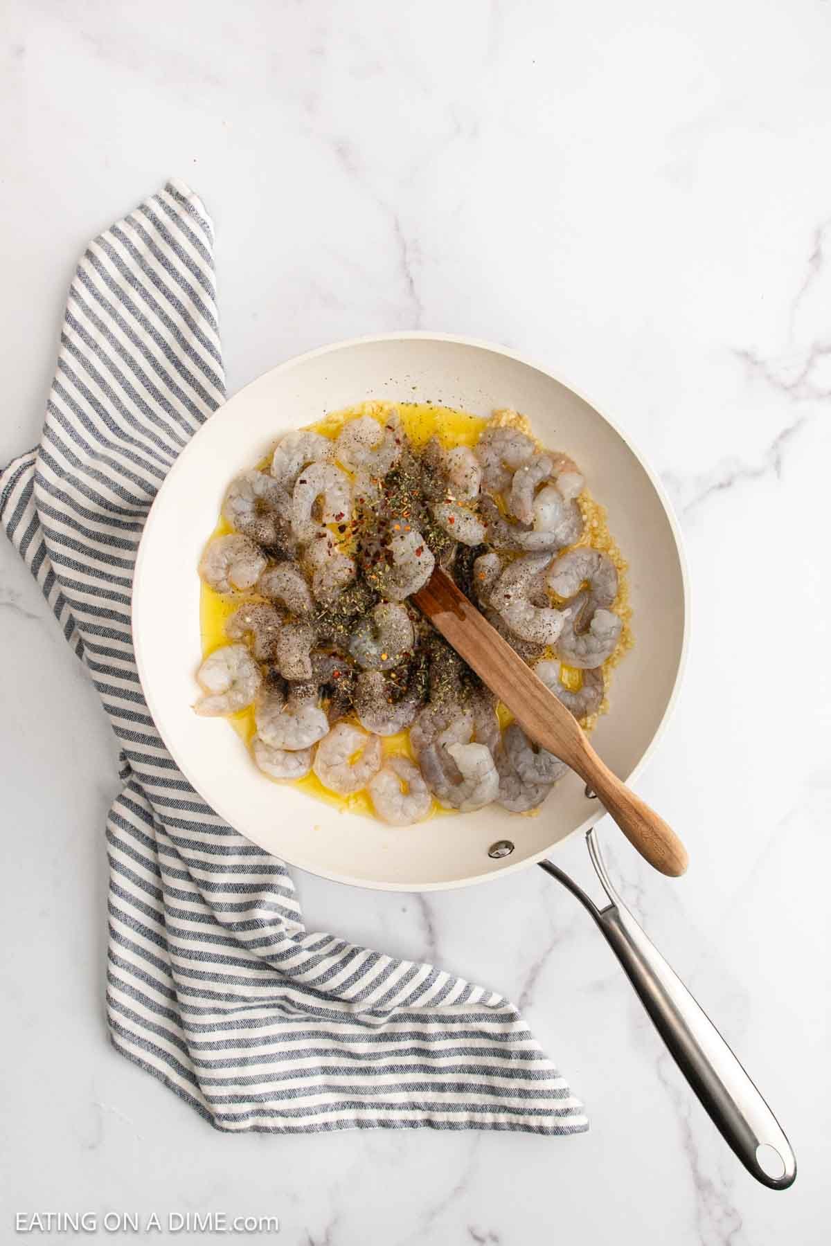 A white skillet with raw shrimp, seasonings, and melted butter for Lemon Garlic Shrimp Pasta is being stirred with a wooden spoon, placed on a striped kitchen towel on a marble countertop.