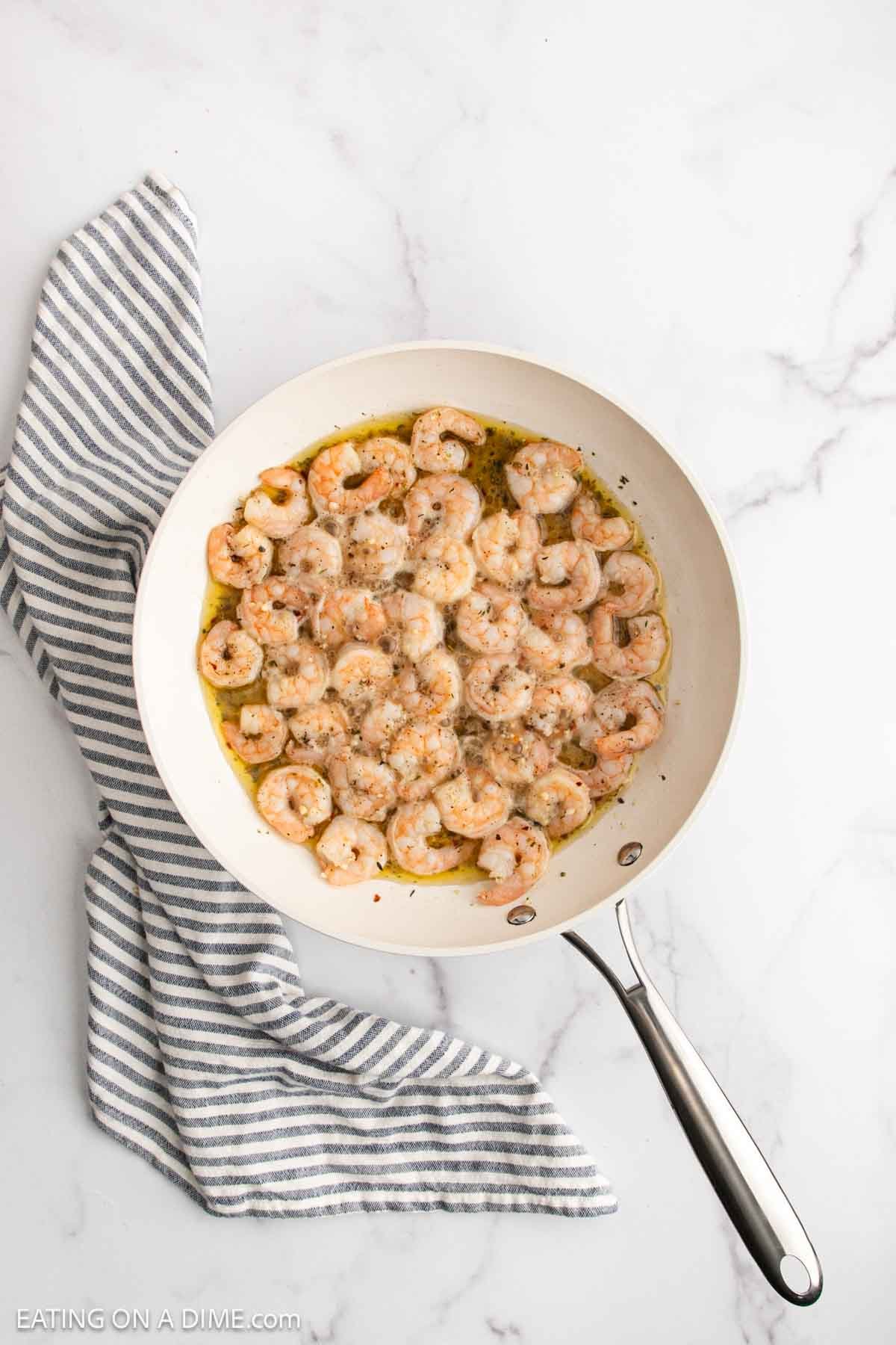 A white skillet filled with Lemon Garlic Shrimp Pasta in a light sauce, garnished with herbs, sits on a marble surface next to a gray and white striped kitchen towel.