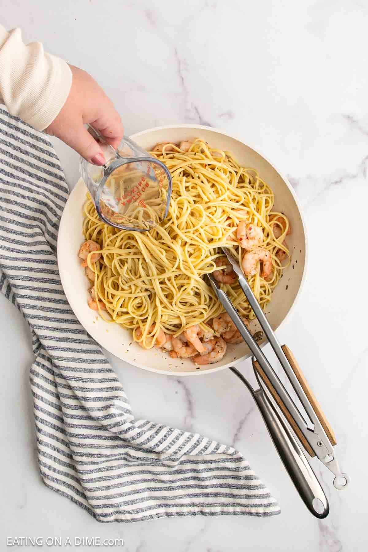A hand pours liquid from a measuring cup into a bowl of Lemon Garlic Shrimp Pasta, with metal tongs and a striped kitchen towel arranged on the marble surface.