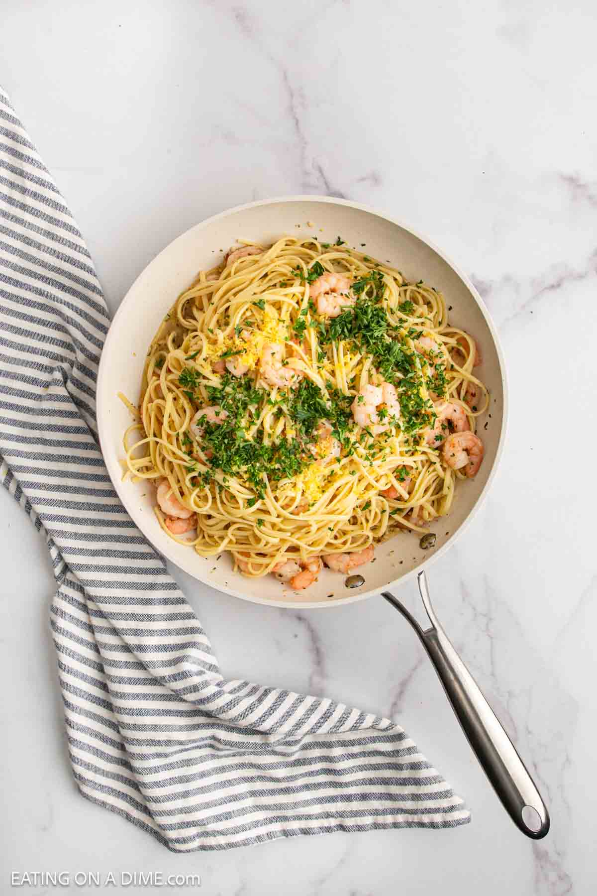 A skillet filled with lemon garlic shrimp pasta, spaghetti, and herbs sits on a marble surface next to a gray and white striped kitchen towel.