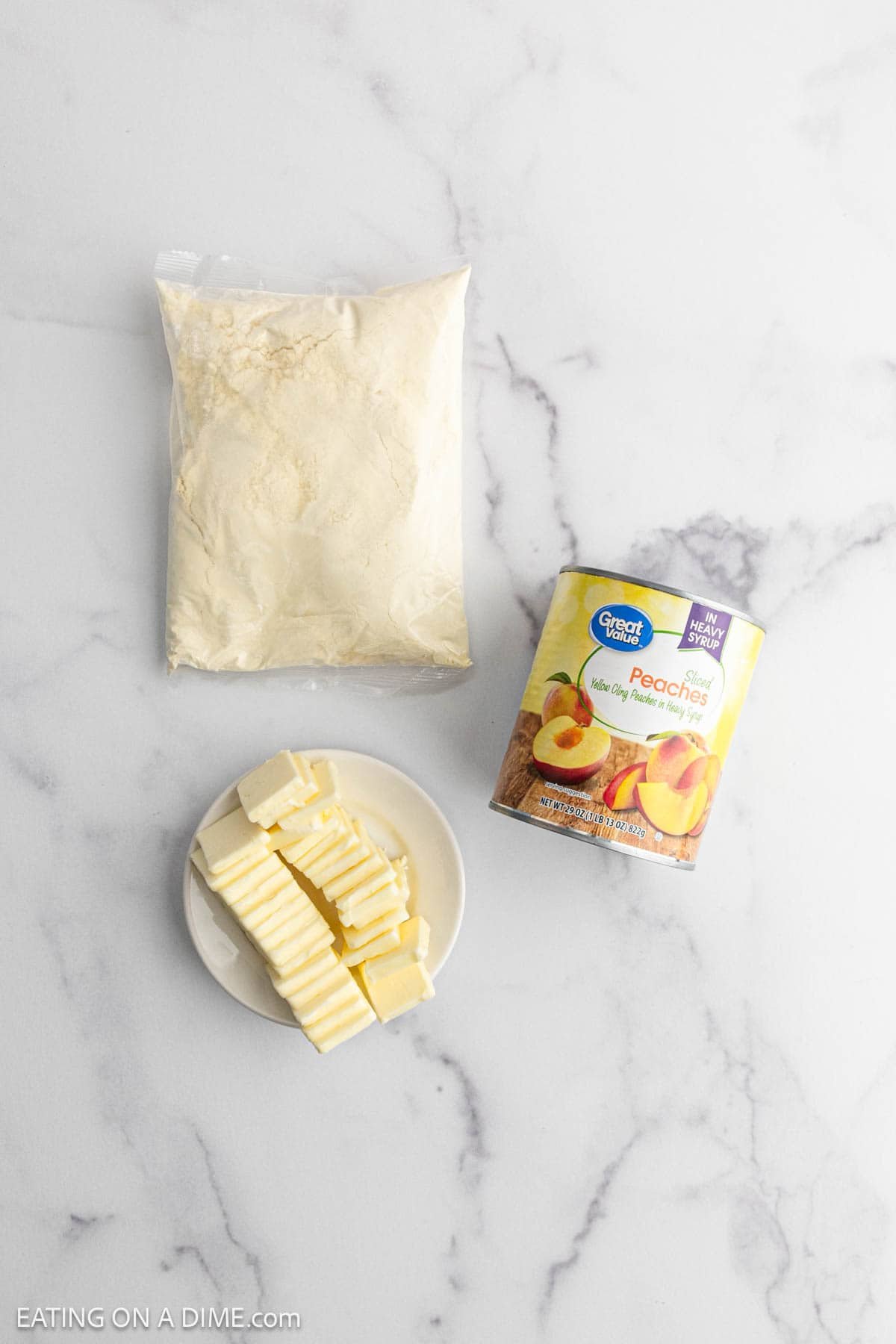 A bag of flour, a can of sliced peaches, and a plate of sliced butter arranged on a white marble surface—perfect ingredients for making a delicious Peach Dump Cake.