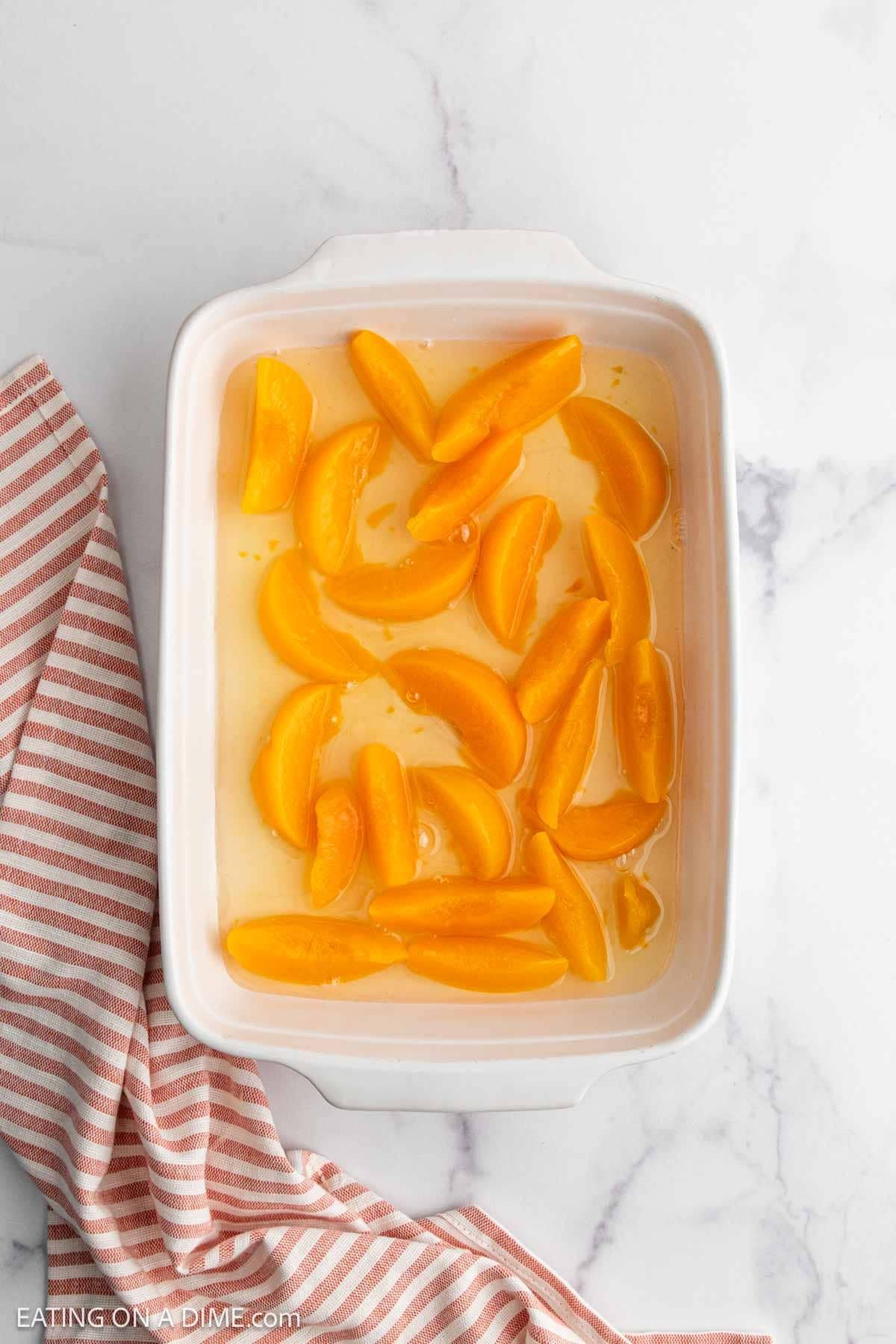 A white baking dish filled with sliced peaches in syrup, ready for a delicious Peach Dump Cake, sits on a marble surface next to a pink and white striped cloth.