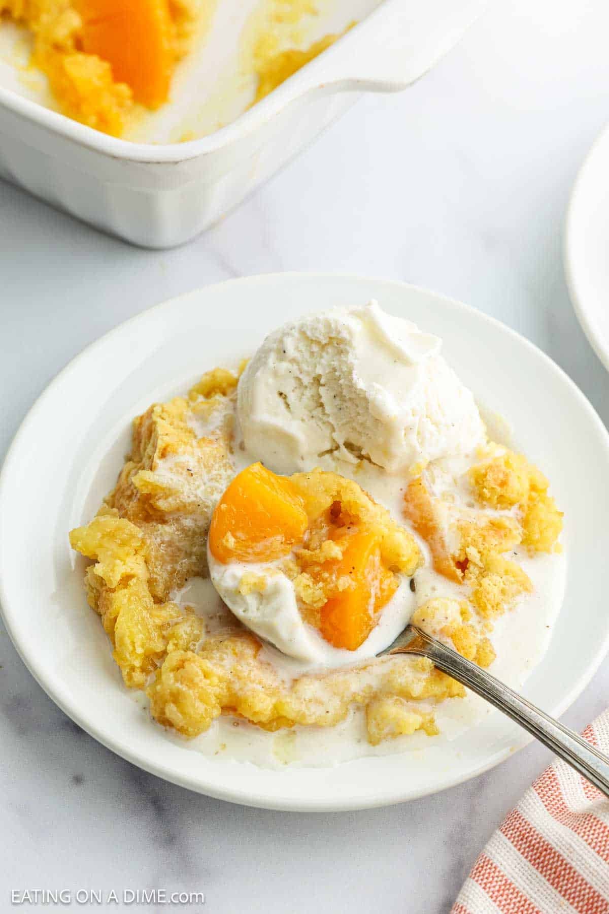 A plate of golden peach cobbler, a classic peach dessert, is topped with melting vanilla ice cream and served with a spoon. Visible peach slices peek through the crust, and a baking dish can be seen in the background.