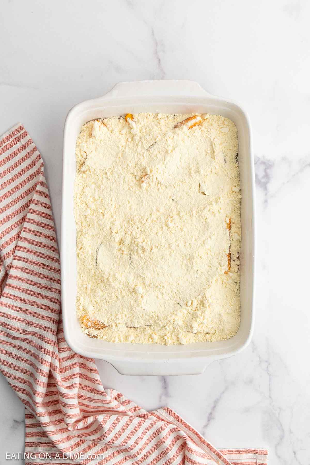 A white baking dish filled with a layer of dry yellow cake mix for Peach Dump Cake, placed on a white marble surface next to a pink and white striped kitchen towel.