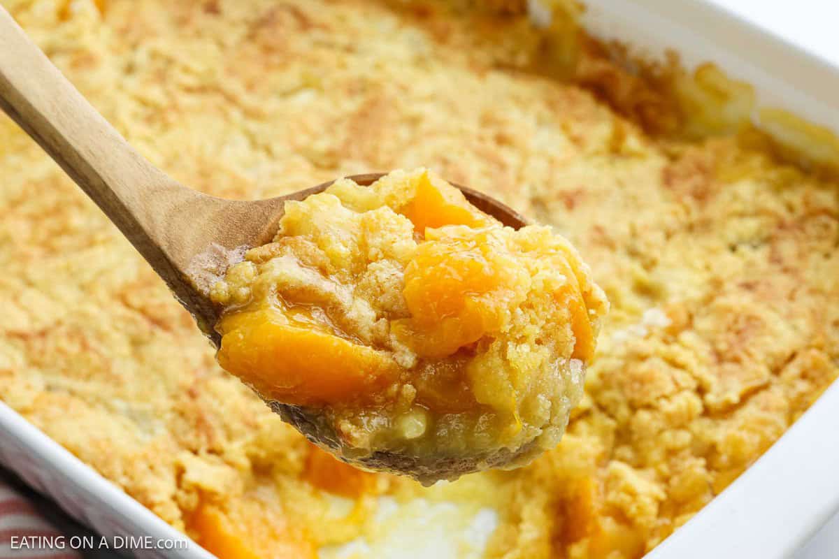 A close-up of a wooden spoon holding a serving of peach dump cake with a golden, crumbly topping, above a baking dish filled with more easy dessert.