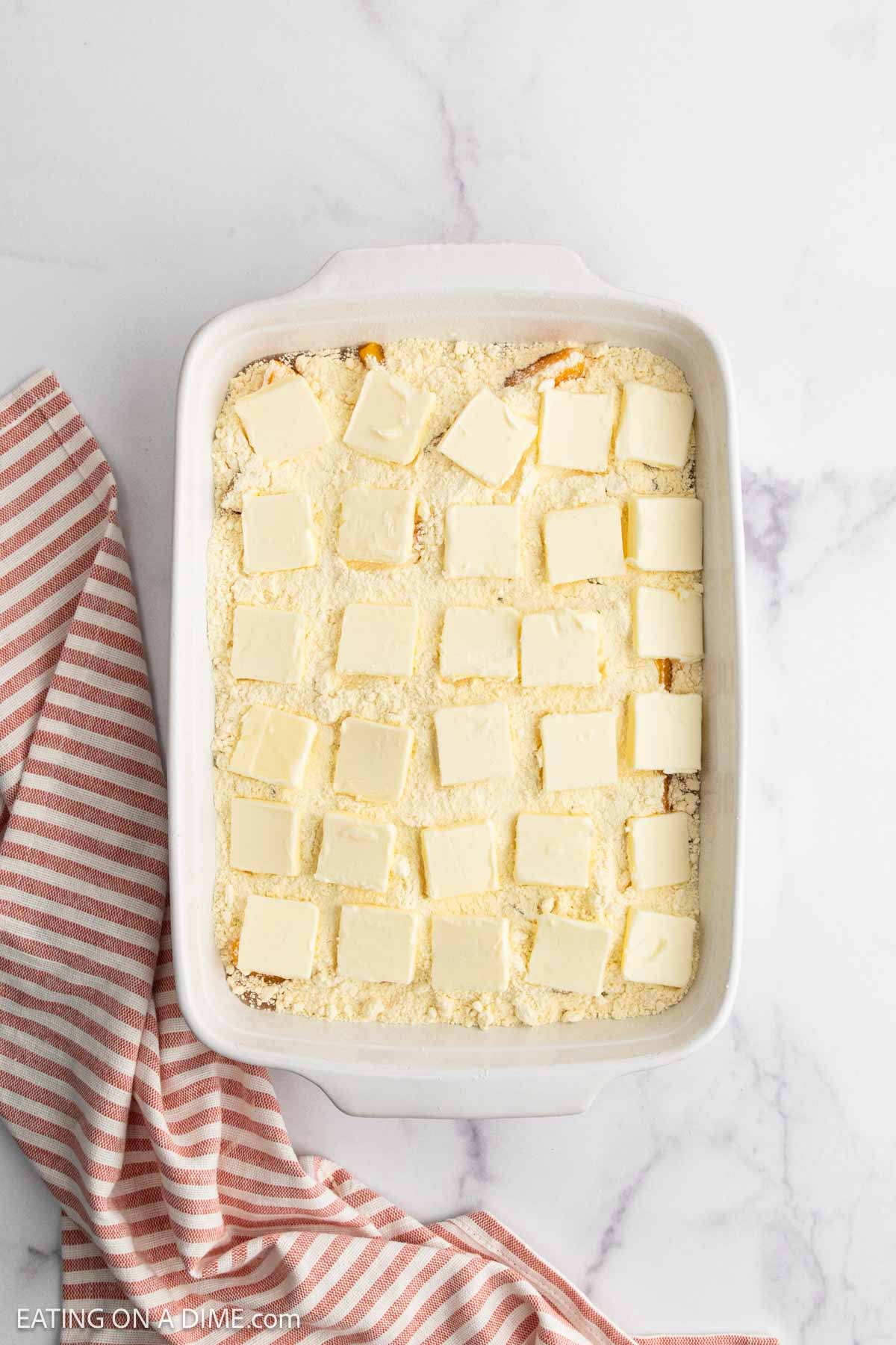A white baking dish filled with an unbaked Peach Dump Cake, topped with evenly spaced square pieces of butter, sits on a white marble surface with a pink-striped kitchen towel nearby.
