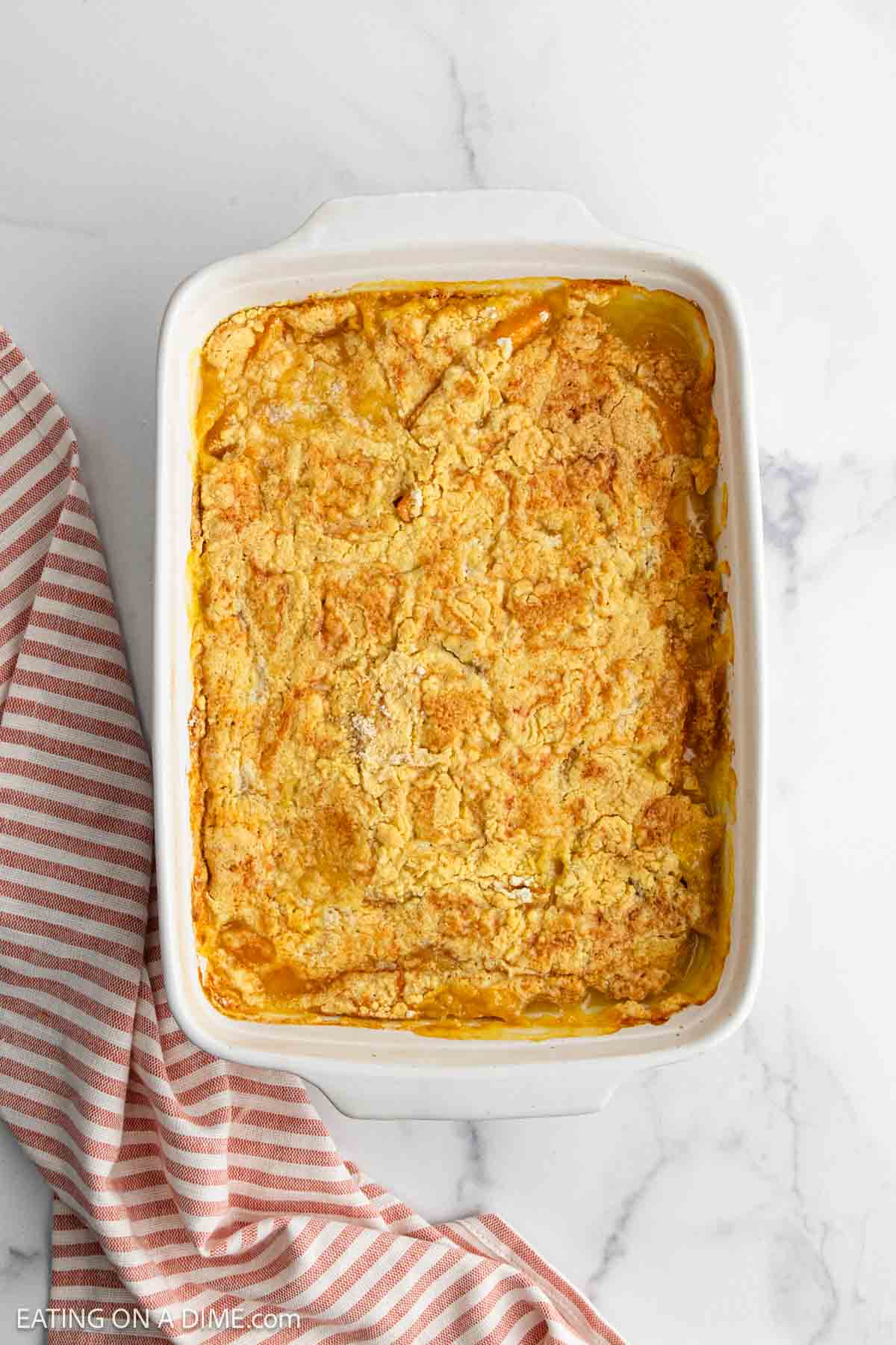 A peach dump cake casserole with a golden, crumbly topping sits in a white dish on a white marble surface, next to a red and white striped cloth.