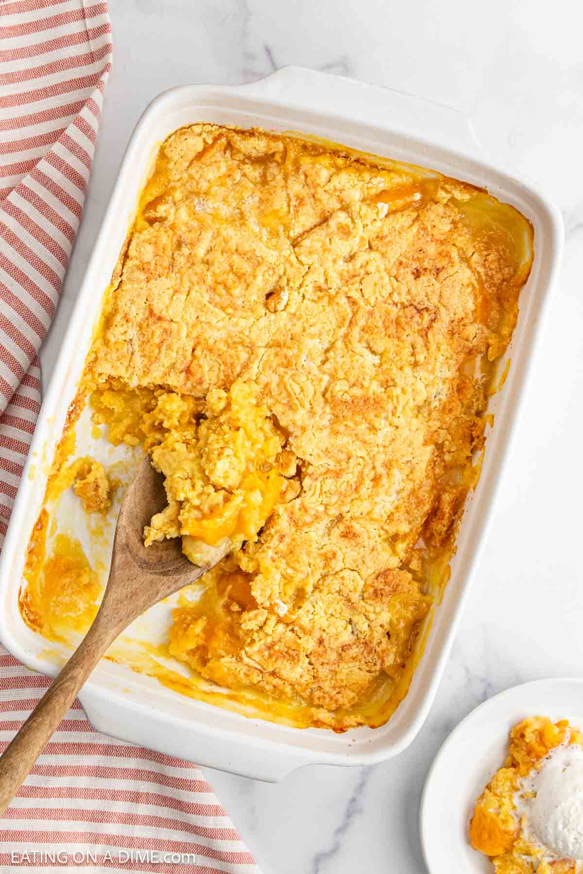 A baking dish filled with golden, crumbly Peach Dump Cake, with a wooden spoon scooping some out. A portion is served on a small white plate nearby, and a striped cloth is next to the dish.