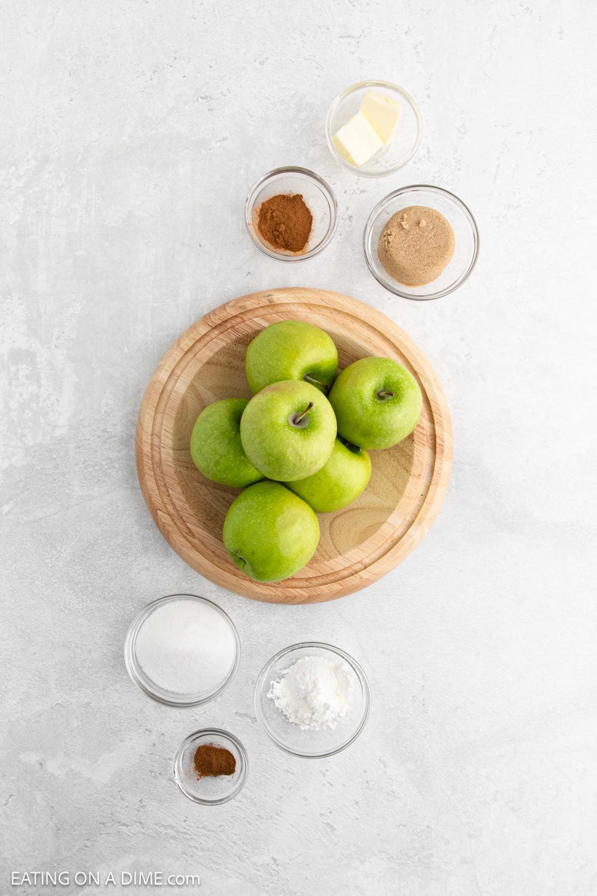 A wooden plate with five green apples sits on a light surface, surrounded by small bowls of butter, brown sugar, cinnamon, white sugar, cornstarch, and ground nutmeg—perfect ingredients for delicious slow cooker apples.