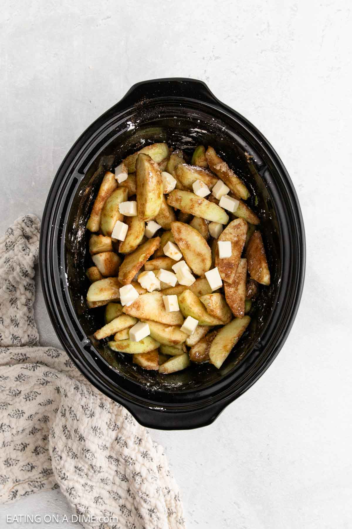 Slow Cooker Apples mixed with cinnamon and topped with butter cubes sit in a black slow cooker, with a gray patterned towel beside it on a light surface.