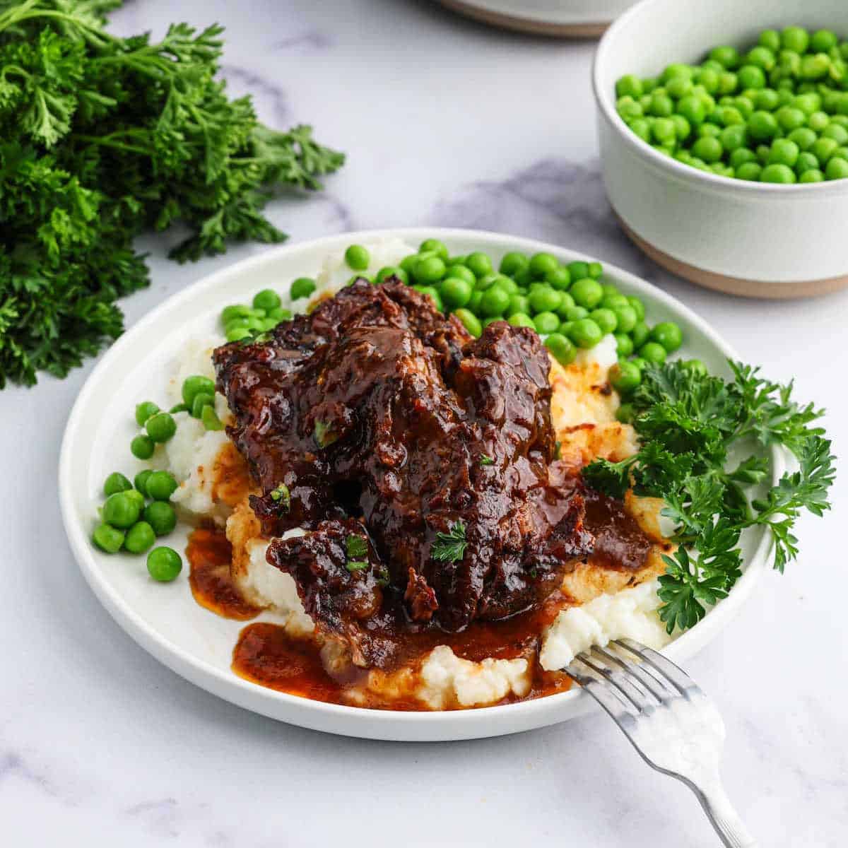 A plate of mashed potatoes topped with Slow Cooker Beef Ribs in rich brown gravy, surrounded by green peas and garnished with fresh parsley. A fork rests on the plate, and parsley bunches are nearby on the table.