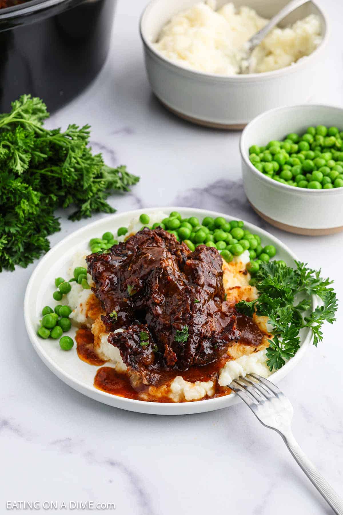 A plate of slow cooker beef ribs with rich brown sauce served over mashed potatoes, garnished with fresh parsley and accompanied by green peas. Bowls of mashed potatoes and peas are in the background.