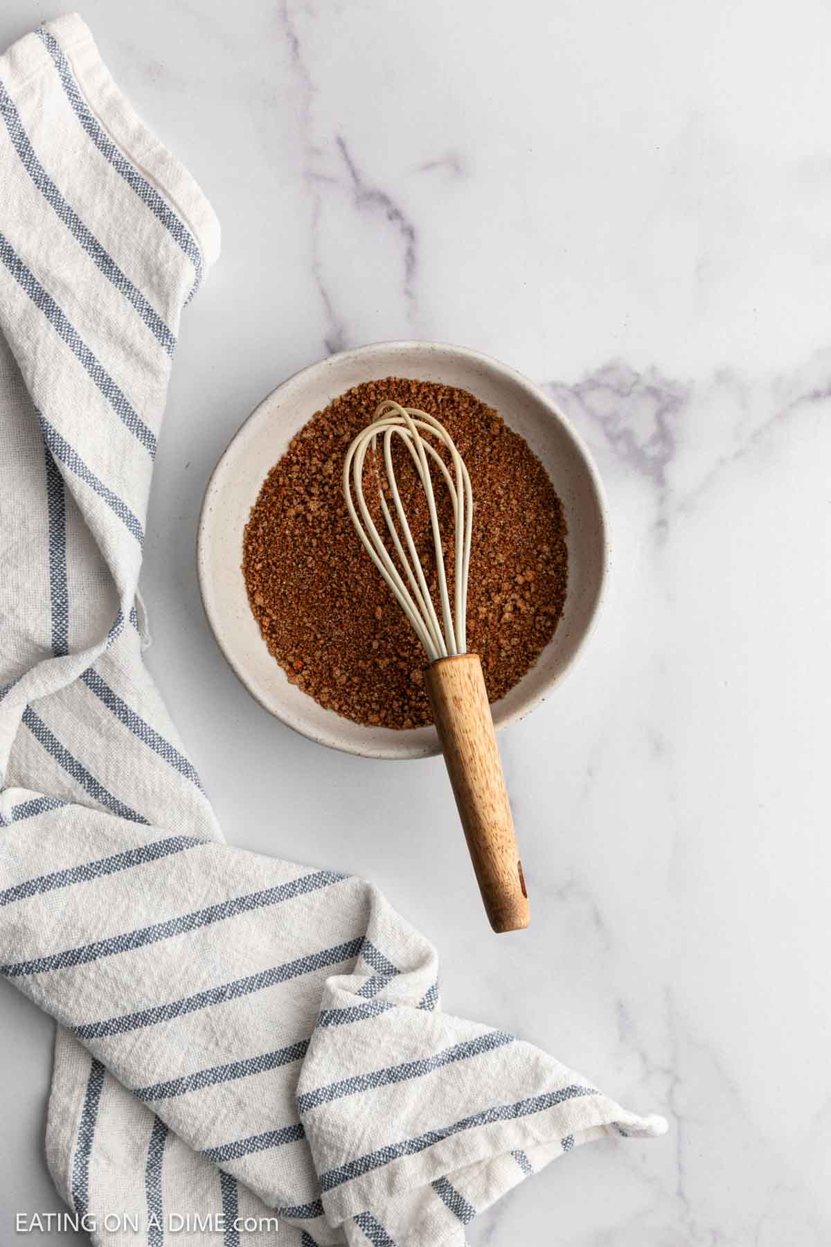 A bowl filled with brown seasoning blend for slow cooker beef ribs and a whisk rests on a marble surface next to a white and blue striped kitchen towel.