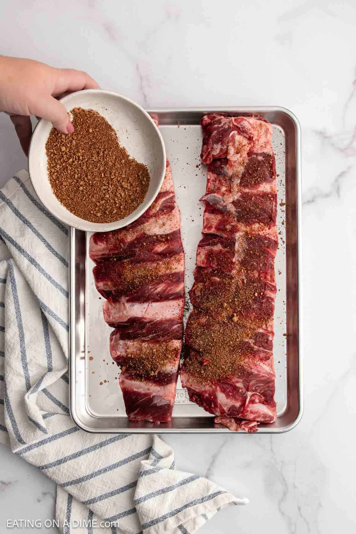 A hand sprinkles brown spice rub from a bowl onto two raw beef ribs on a metal tray, hinting at flavorful Slow Cooker Beef Ribs, with a striped towel beside them on a marble countertop.