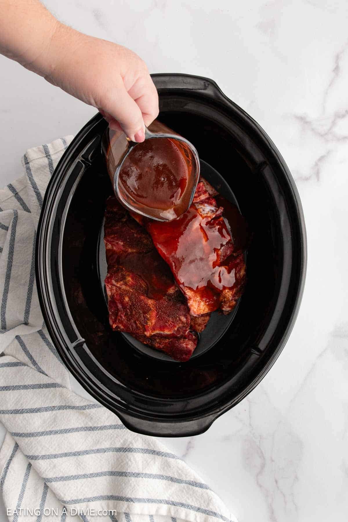 A hand pours barbecue sauce from a glass jar over seasoned beef ribs in a black slow cooker, with a striped kitchen towel beside it on a white marble surface.