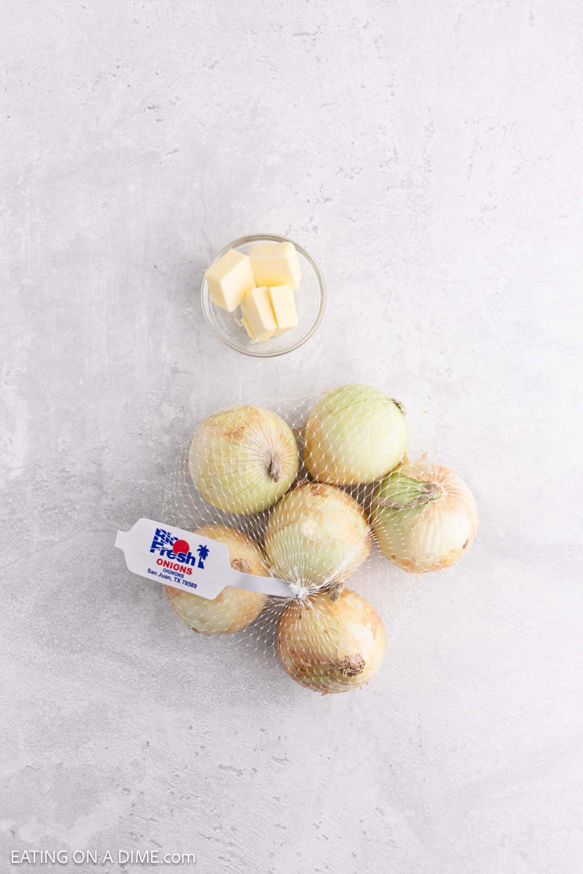 A small glass bowl with butter cubes sits next to a mesh bag filled with yellow onions, perfect for making caramelized onions, on a light gray surface. The bag has a white label with blue and red text.