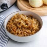 A white bowl filled with caramelized onions sits on a light surface next to a blue-striped towel. In the background, whole onions rest on a cutting board beside a slow cooker, hinting at the savory process behind this dish.