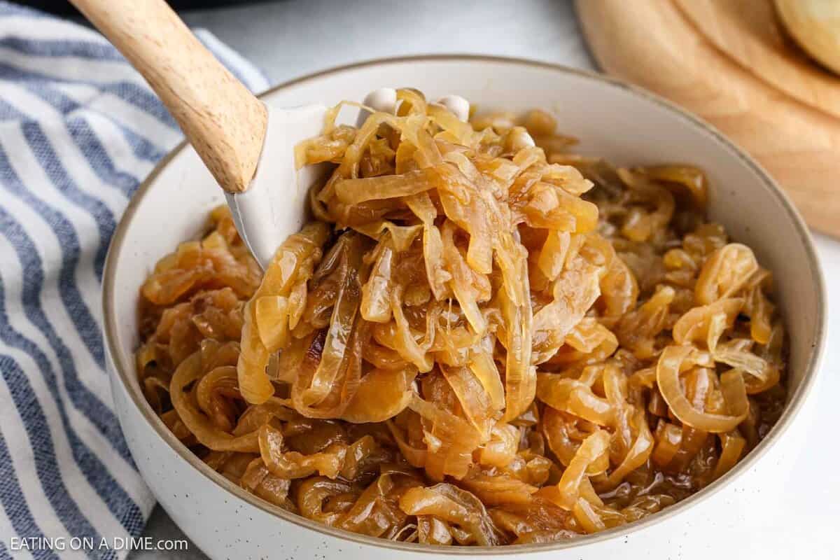 A bowl of slow cooker caramelized onions with a wooden spoon lifting some up, showing the soft, golden-brown onions. A striped cloth is partially visible beside the bowl.