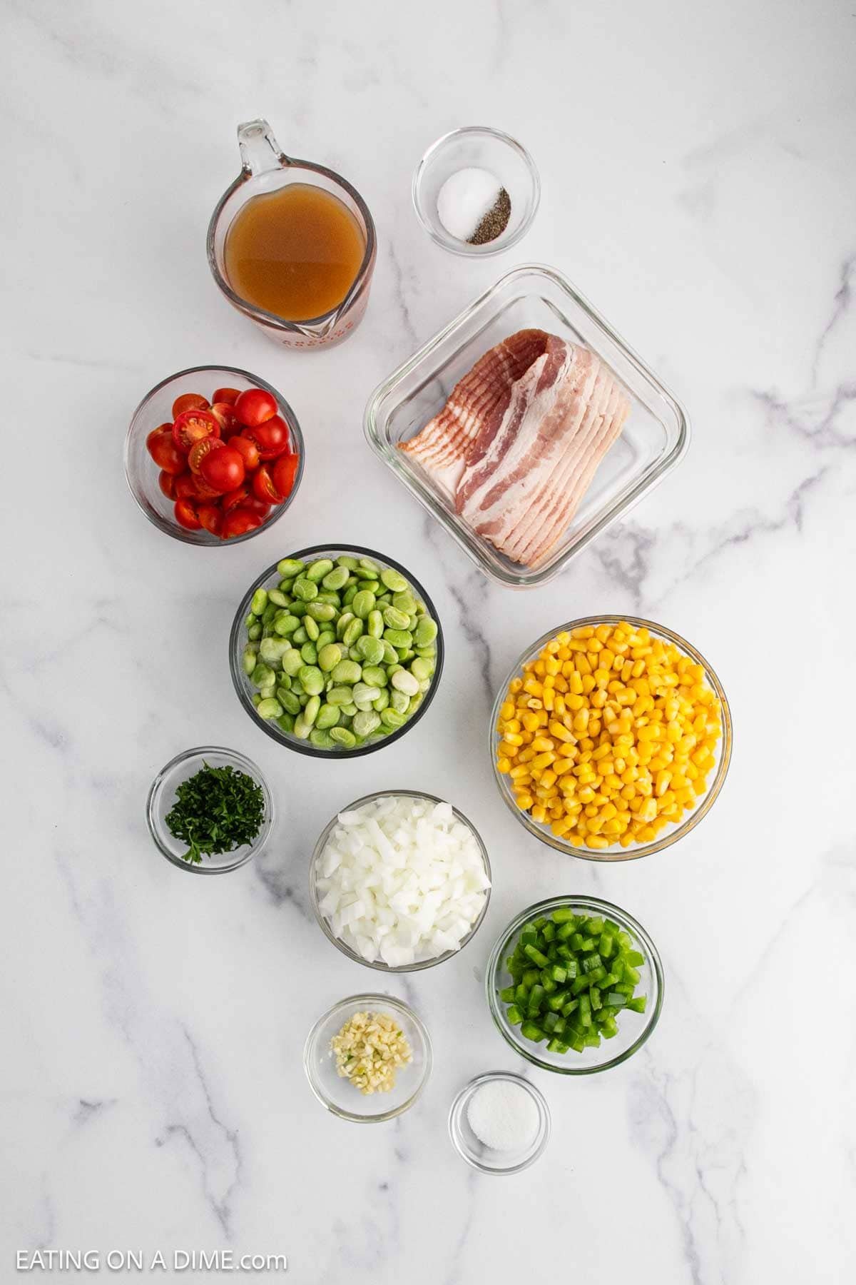 Overhead view of ingredients in bowls on a marble surface: bacon, cherry tomatoes, lima beans, corn, chopped onion, green bell pepper, minced garlic, chopped herbs, salt, pepper, and broth.