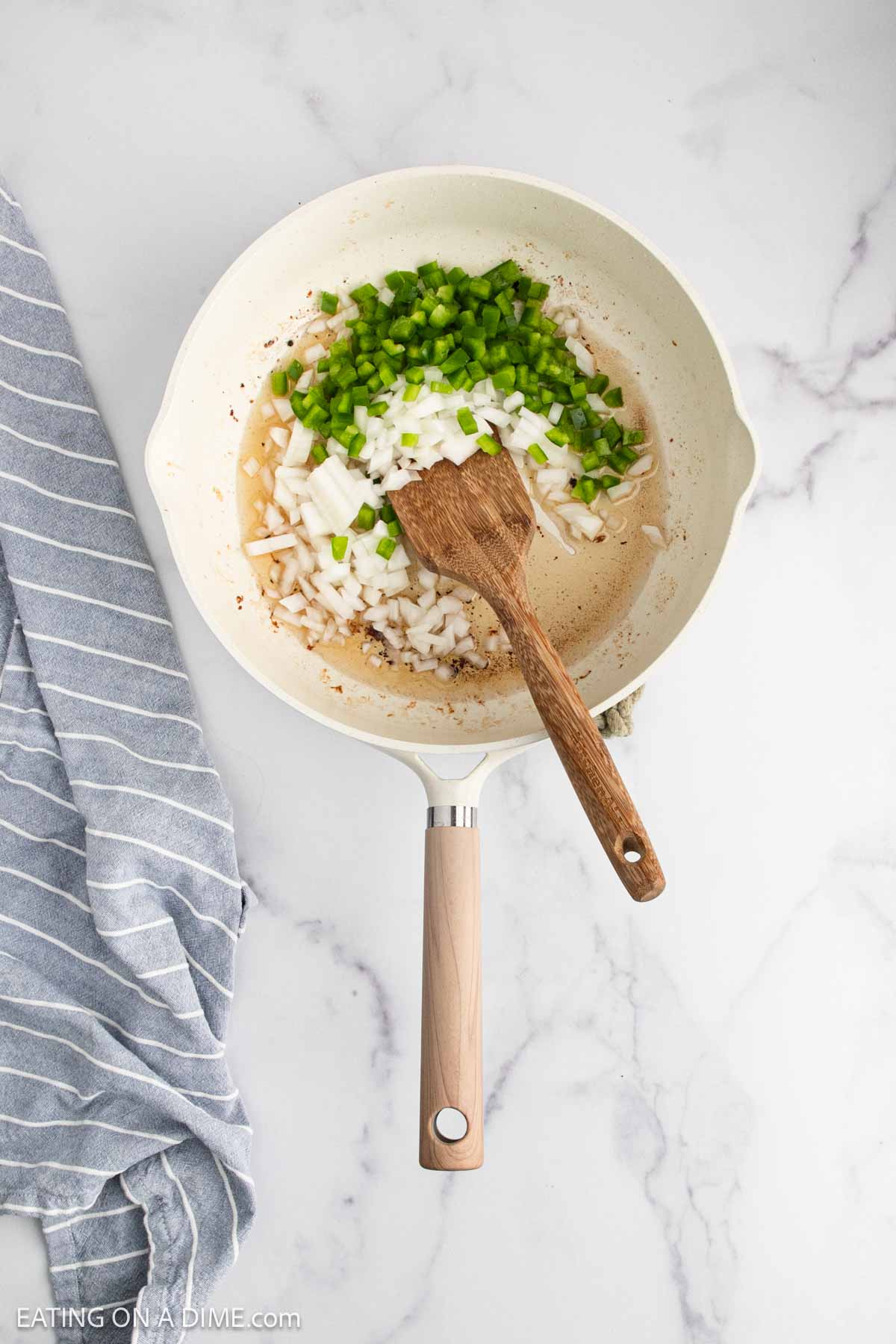 A skillet on a marble countertop contains chopped onions and green bell peppers being sautéed with a wooden spatula. A gray and white striped kitchen towel is beside the skillet.