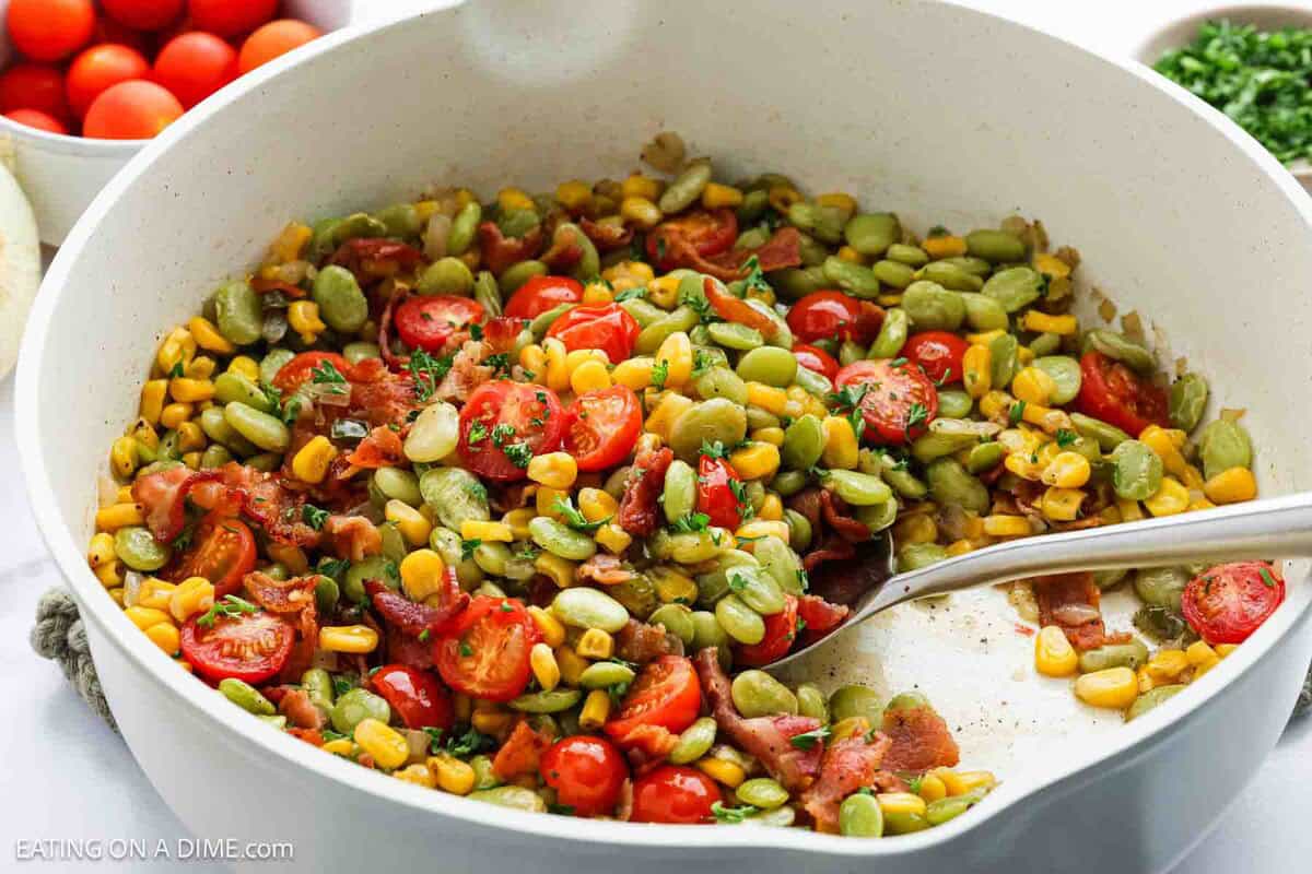 A white bowl filled with a colorful salad made of corn, lima beans, cherry tomatoes, chopped bacon, and herbs, with a spoon resting inside. Bowls of cherry tomatoes and greens are in the background.