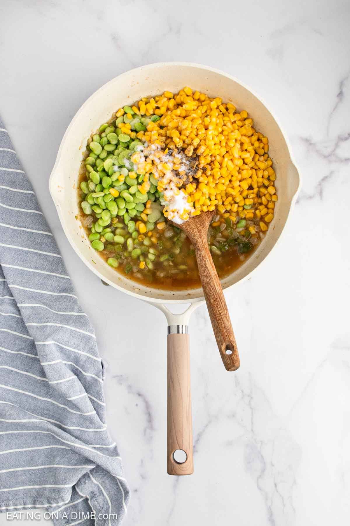 A white skillet with corn, lima beans, chopped vegetables, and seasonings being mixed with a wooden spoon, sits on a marble countertop next to a gray and white striped towel.