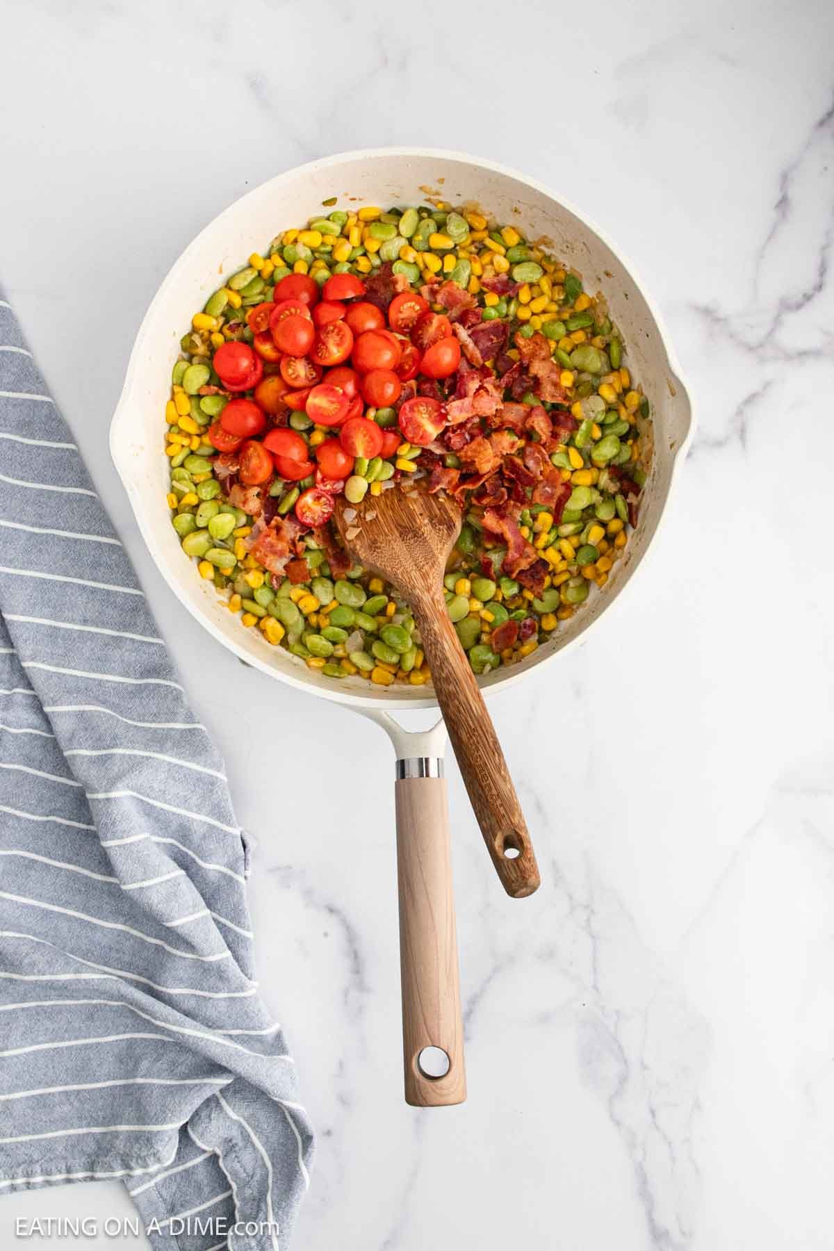 A skillet containing a colorful mixture of corn, lima beans, cherry tomatoes, and crumbled bacon sits on a marble surface next to a gray and white striped towel.