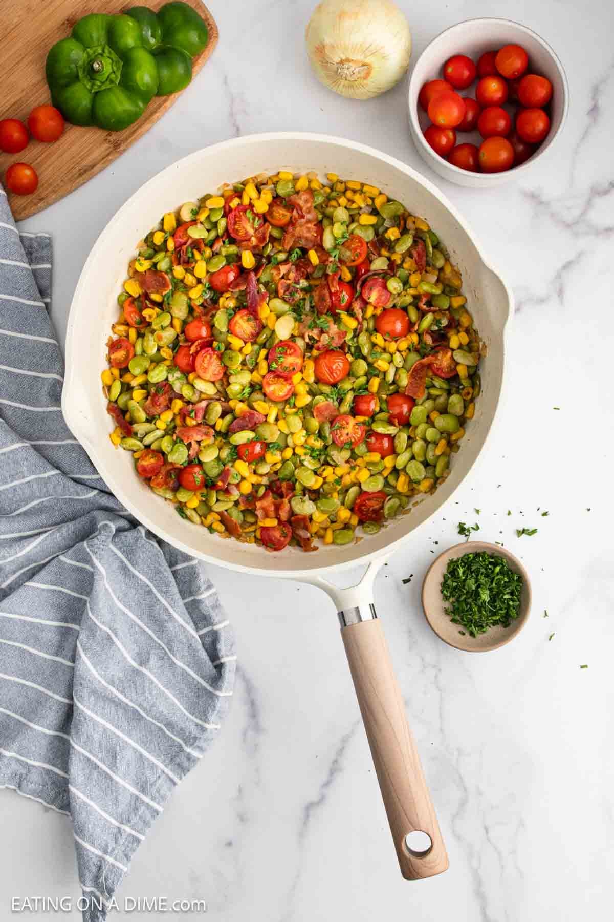A skillet of succotash containing corn, lima beans, and cherry tomatoes sits on a marble countertop. Surrounding the skillet are fresh cherry tomatoes, a green bell pepper, an onion, chopped herbs, and a striped towel.