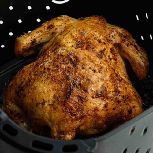 An Air Fryer Whole Chicken with golden, crispy skin sits in a black air fryer basket, seasoned generously with visible herbs and spices.