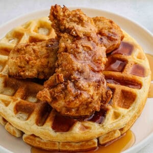 A close-up of crispy chicken and waffles features golden fried chicken tenders atop a fluffy waffle, drizzled with syrup and served on a white plate.