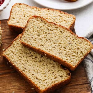 Three thick slices of homemade gluten-free Cottage Cheese Bread rest on a wooden cutting board, showing a dense, lightly textured crumb and golden brown crust. A white plate with more bread is visible in the background.
