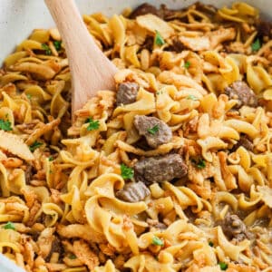 A close-up of French Onion Beef and Noodles mixed in a creamy sauce, garnished with chopped parsley, being stirred with a wooden spoon.